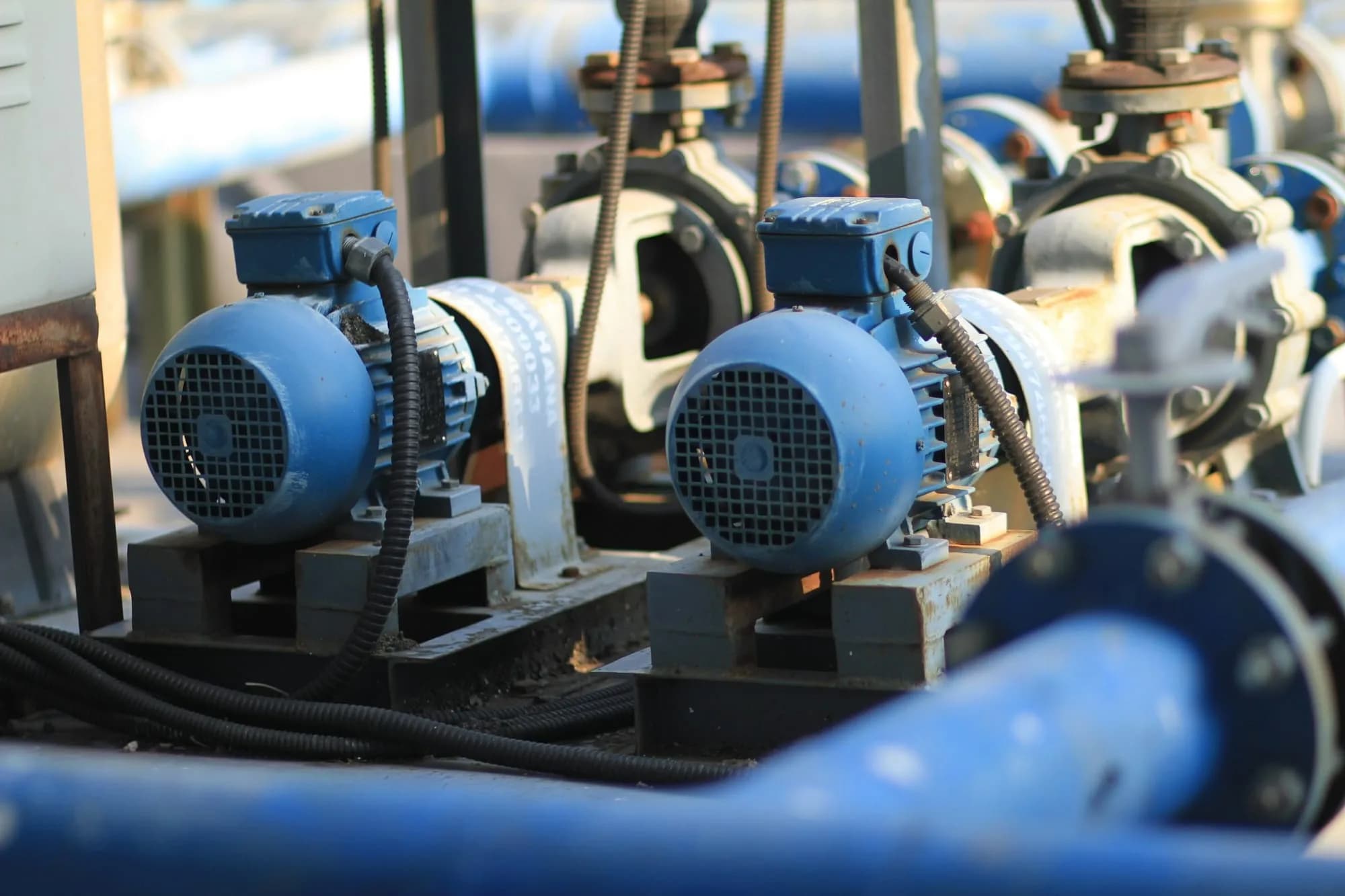 Dos motores eléctricos azules acoplados a bombas sobre su base, conectados a cañerías y válvulas en una sala de máquinas
