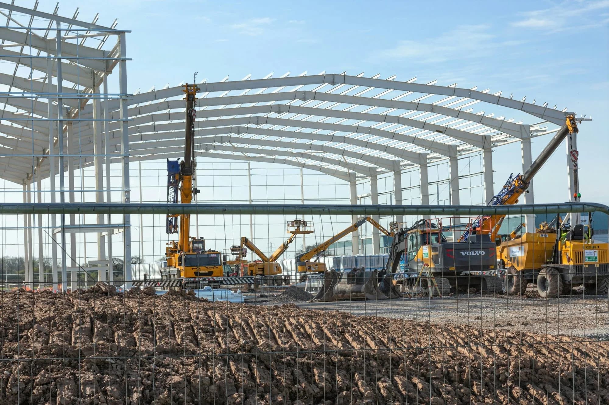 Estructura de acero de un galpón en construcción, con grúas, excavadoras y maquinaria de obra en primer plano
